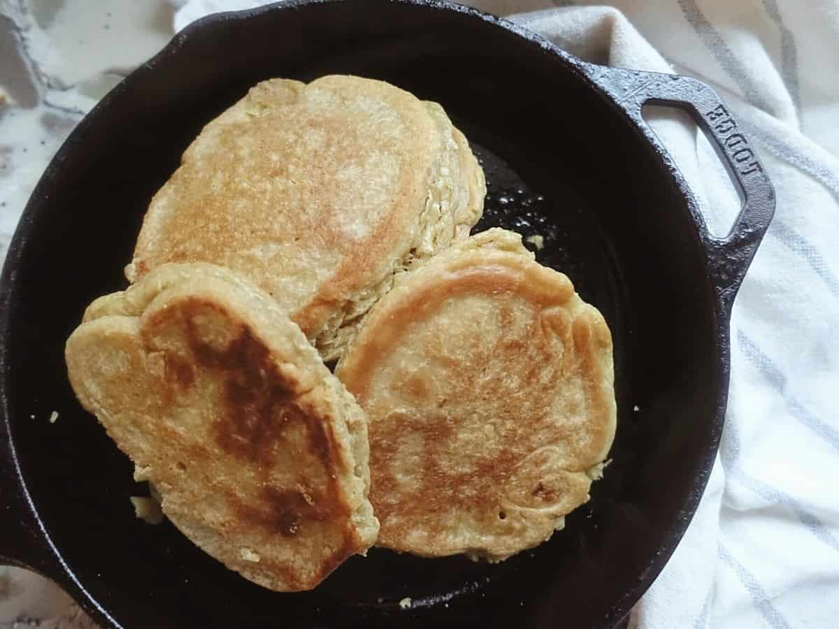 thick sourdough pancakes in a cast iron skillet with a blue and white striped tea towel in the back
