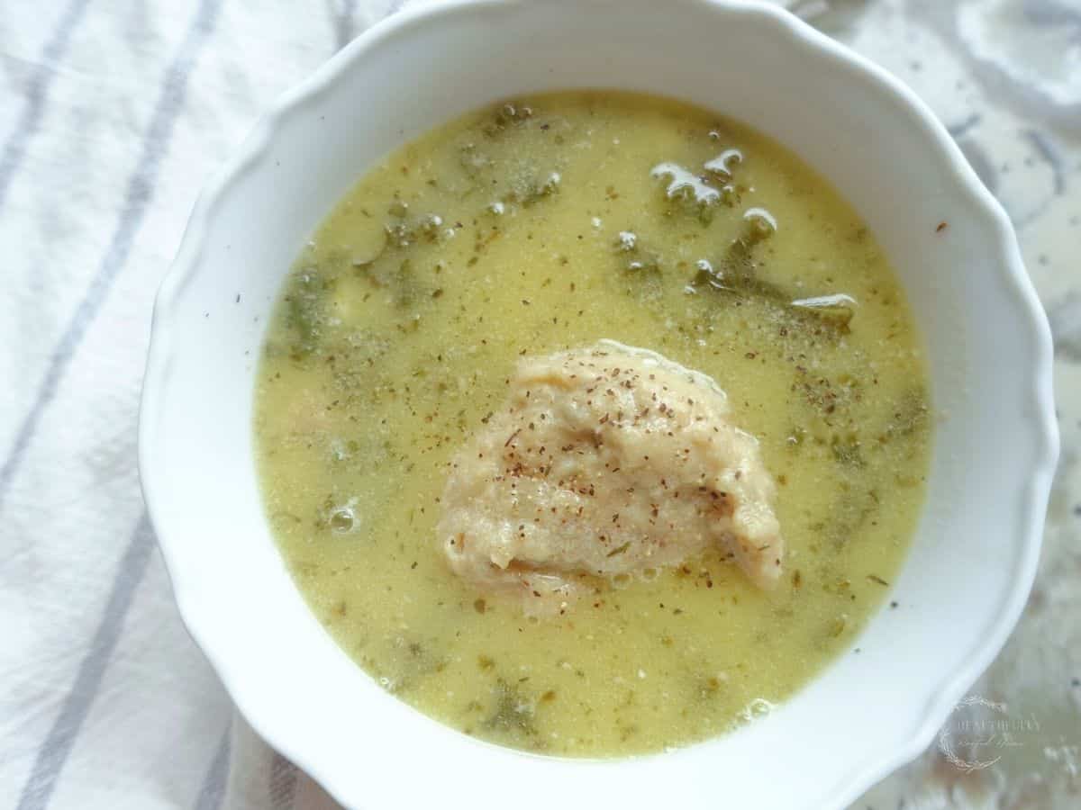 kale chicken soup with a sourdough dumpling in a white bowl and a blue and white striped tea towel on the side