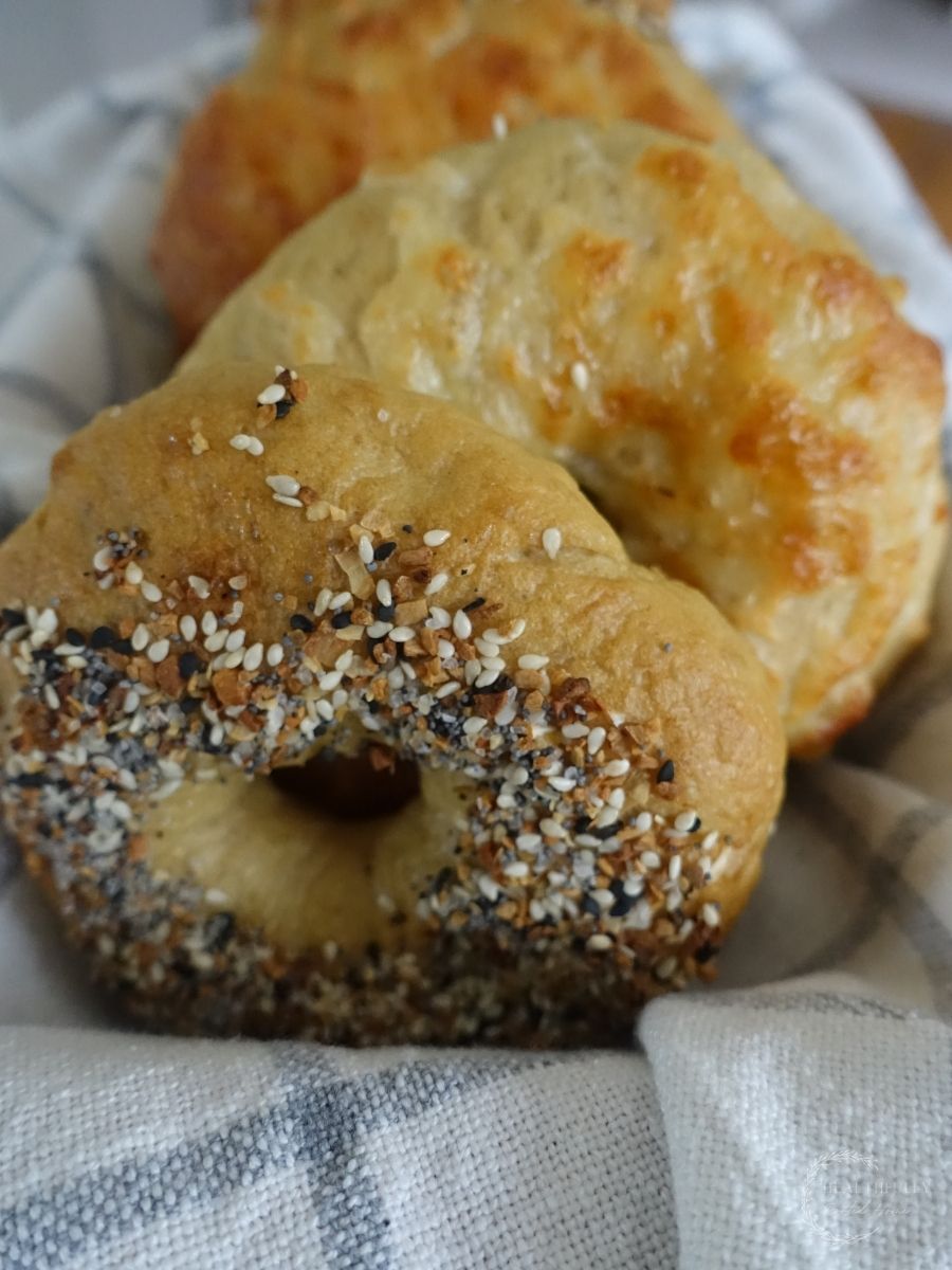 Sourdough discard bagels in a basket lined with a ticking striped tea towel showing off the bagel toppings of everything bagel seasoning and cheddar cheese