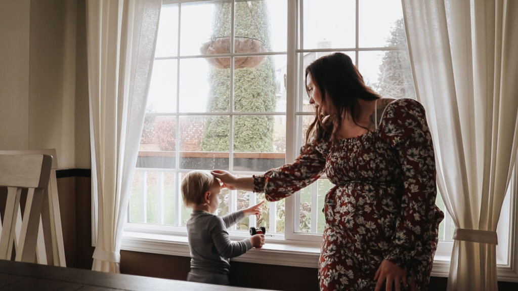 christian homemaker sitting on a windowsill playing with son's hair while he plays with trucks and looks out the window