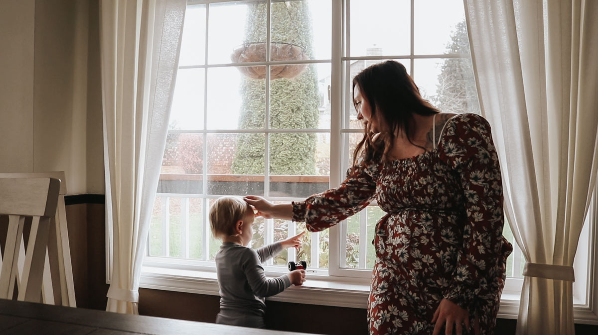 christian homemaker sitting on a windowsill playing with son's hair while he plays with trucks and looks out the window