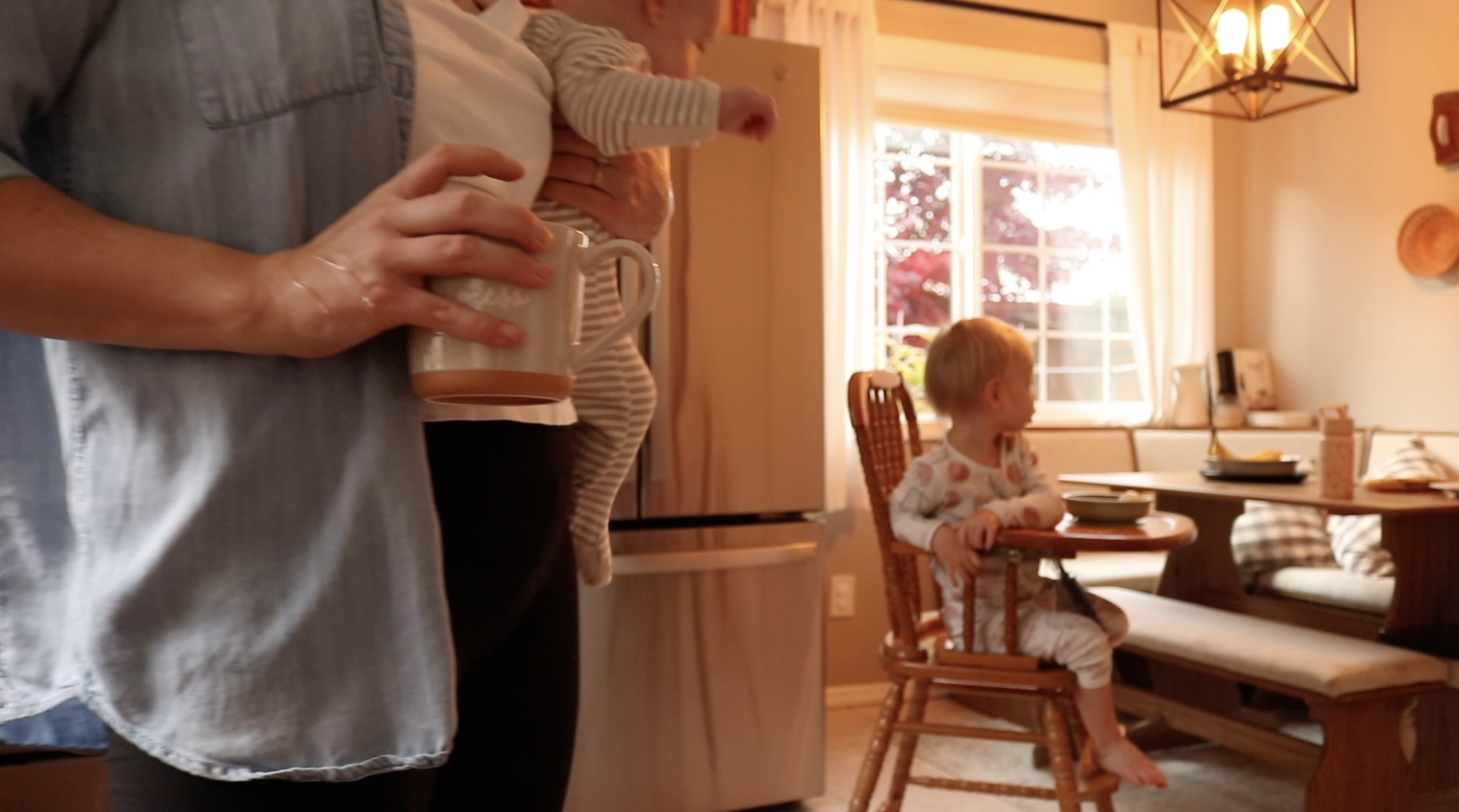 mom holding a baby and coffee with another child in the background in a high chair