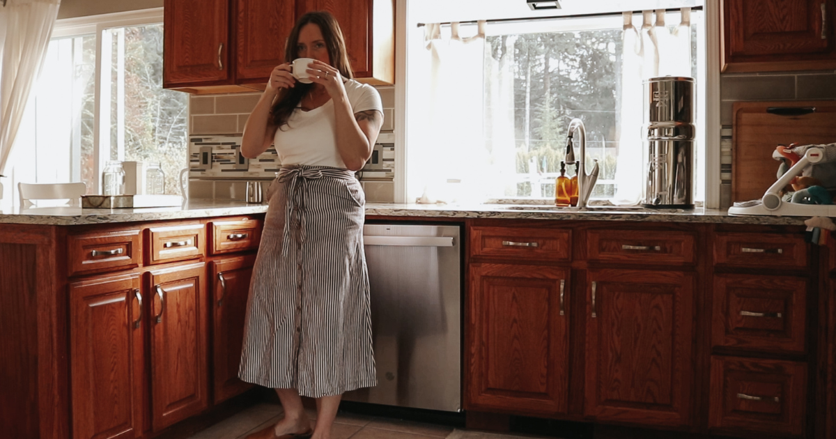 living slowly as a christian homemaker shes drinking her coffee in a long skirt in the kitchen