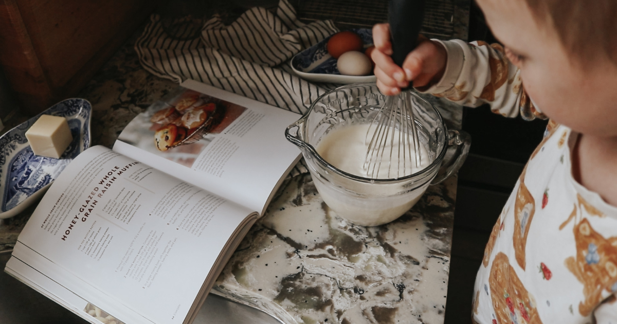 Boy stands at kitchen counter whisking muffin batter while a cook book sits open to a muffin recipe on the counter