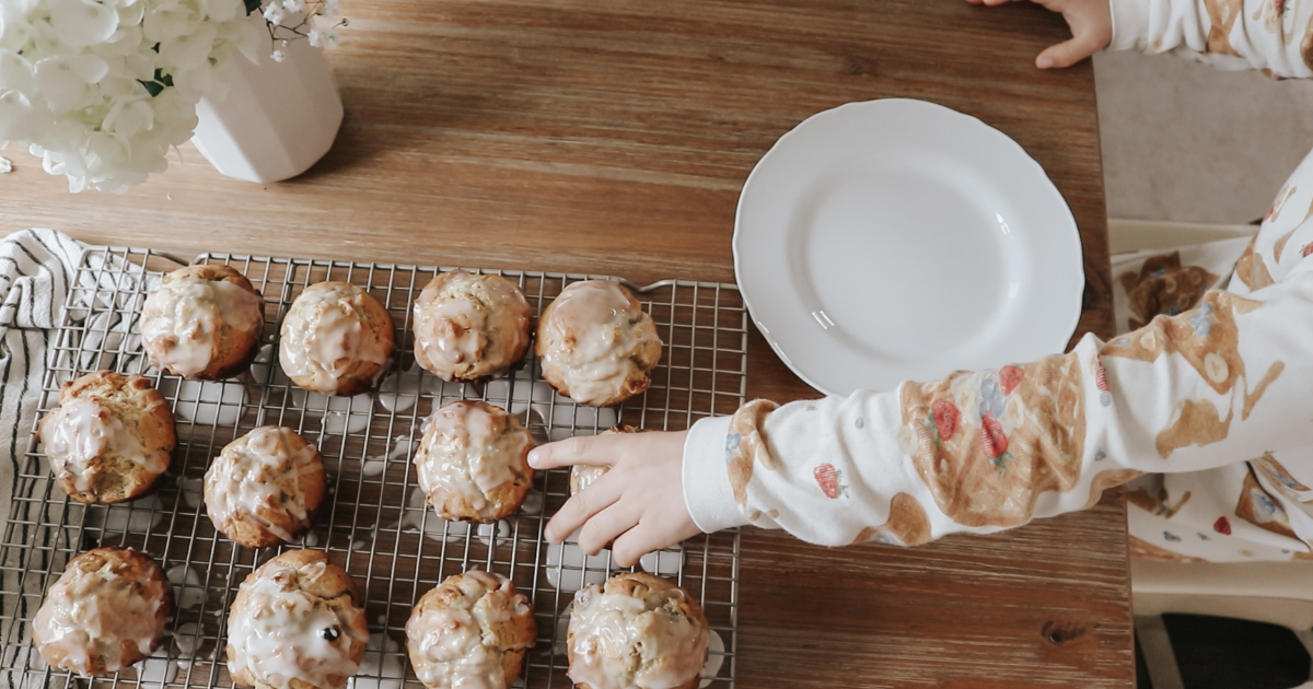 A small hand reaches for a muffin that is baked, iced, and cooling on a rack
