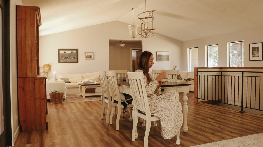 Christian homemaker wearing a floral dress sits at her dining room table where she is eating a bowl of oatmeal
