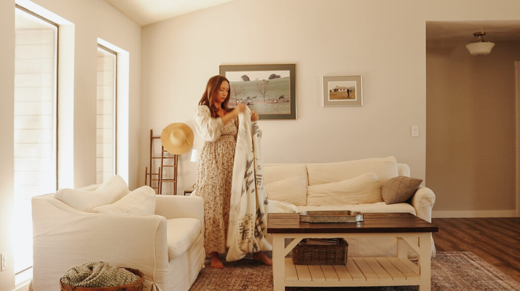 Christian homemaker wearing a floral dress stands in her living room next to the couch holding a blanket that she is going to fold