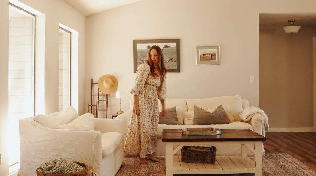 Christian homemaker wearing a floral dress stands next to the white couch in her living room