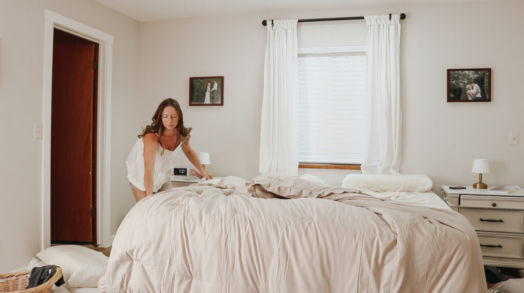Christian homemaker wearing white tank top and denim shorts is making her bed.