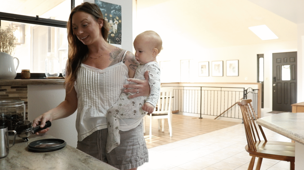 Christian homemaker wearing a white tank top and denim shorts holds a baby on her hip while making coffee at the kitchen counter.