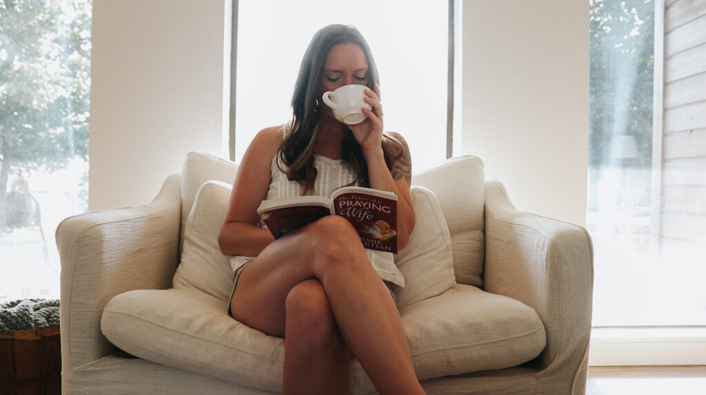 Christian homemaker wearing a white tank top and denim shorts is sitting on her couch while reading a book and drinking a cup of coffee.