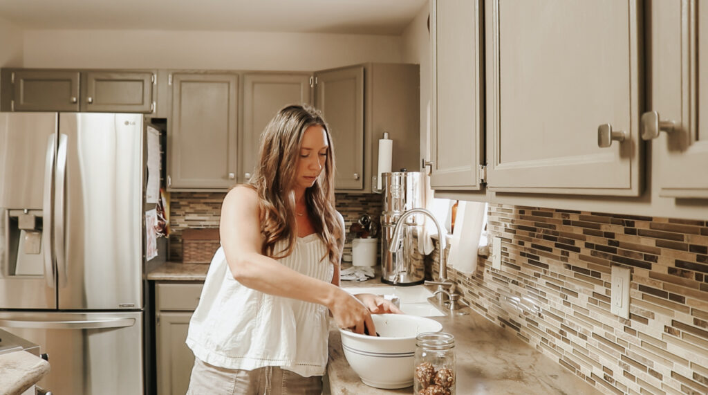 Christian homemaker wearing a white tank top and denim shorts is standing at her kitchen counter scooping dough out of a bowl into a container.