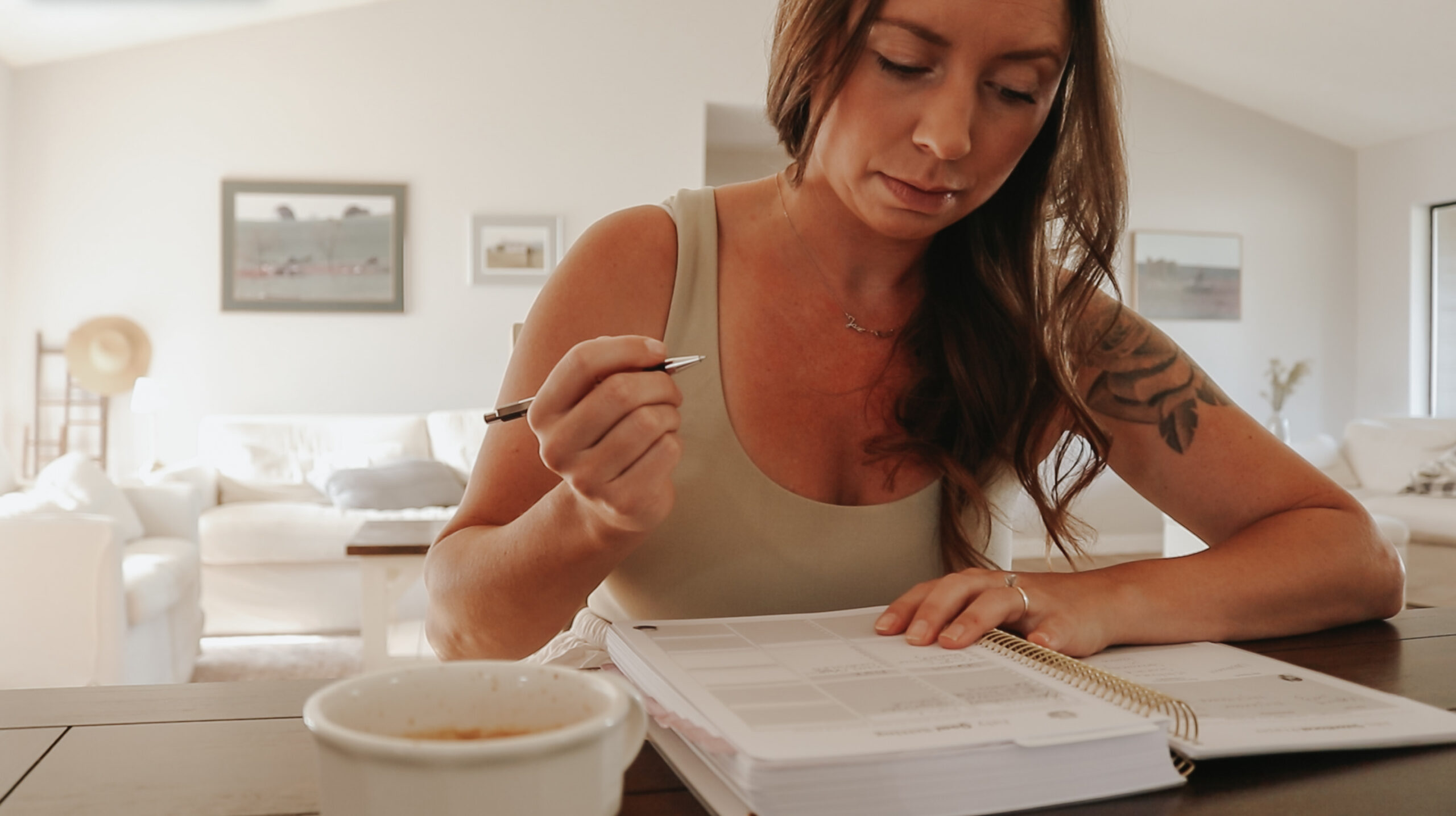 Christian homemaker wearing a cream colored tank top is sitting down at her kitchen table with a cup of coffee and is writing in a notebook.