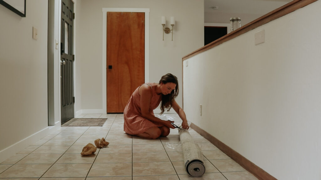 Christian homemaker wearing a peach colored dress is cutting the plastic on a brand new rolled up rug while sitting on the floor of her entryway.