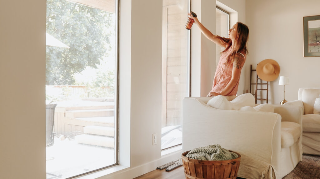 Christian homemaker wearing a peach colored dress is spraying her living room windows with cleaning spray.