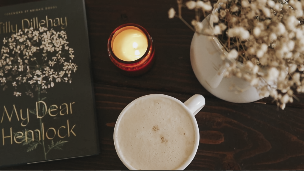 A coffee mug filled with coffee and frothed milk on top sits next to a book titled "My Dear Hemlock" on a table.