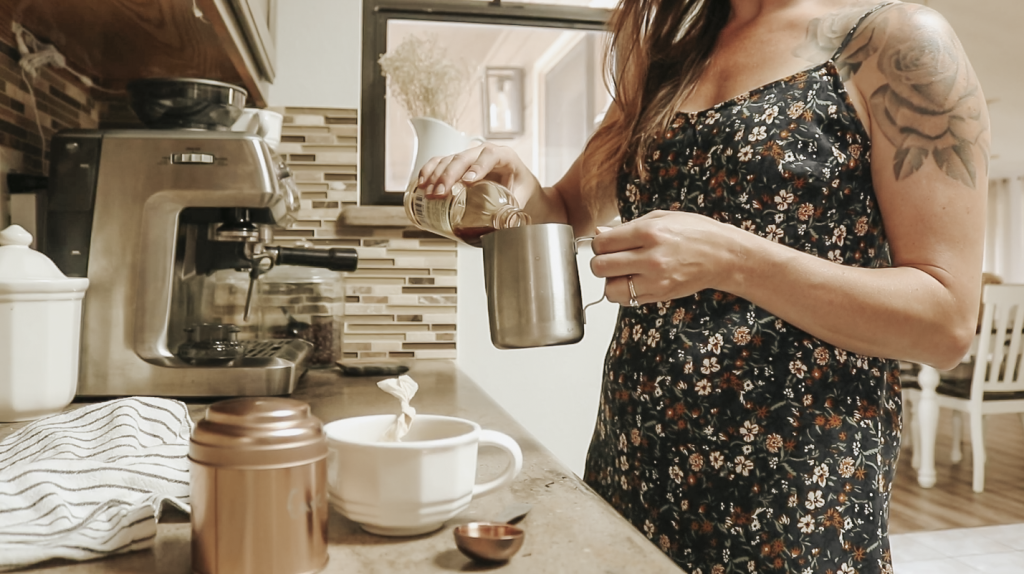 Christian homemaker wearing a floral dress pours milk into a metal frothing cup beside a coffee machine on a counter.