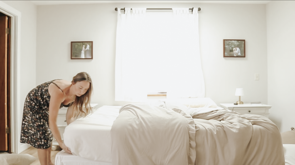 Christian homemaker wearing a black floral dress puts a clean white fitted sheet onto her bed.
