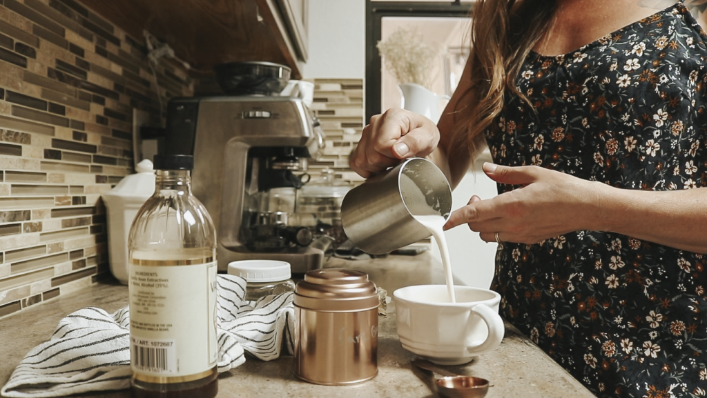 Christian homemaker wearing a floral dress pours frothed milk into a coffee mug on the counter.