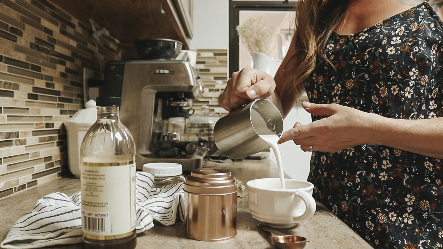Christian homemaker wearing a floral dress pours frothed milk into a coffee mug on the counter.