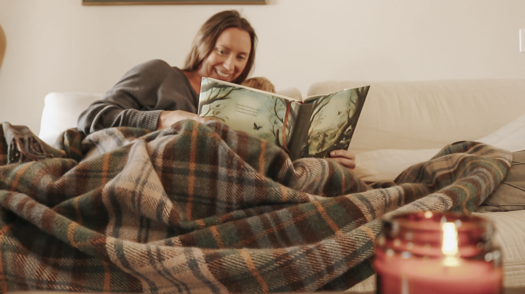 Christian homemaker sitting on a couch reading a picture to a book to her child under a plaid blanket.