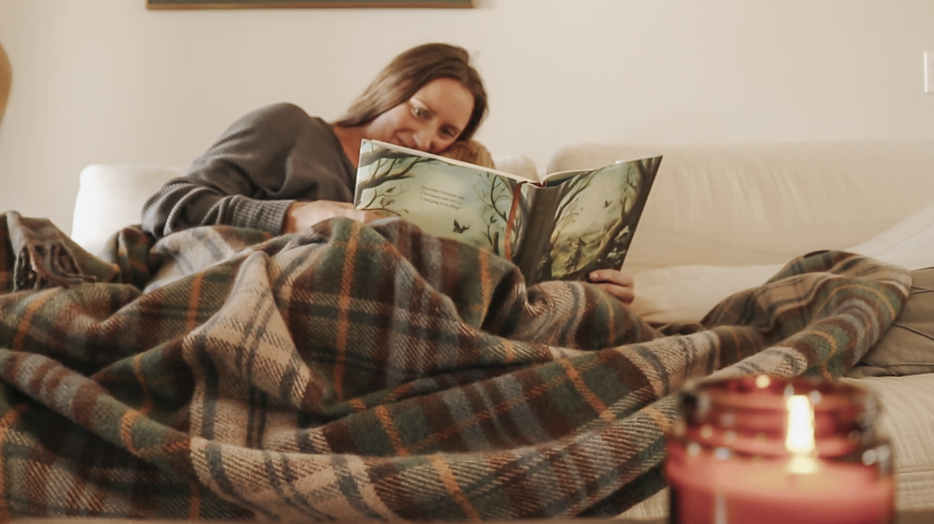 Christian homemaker sitting on a couch reading a picture to a book to her child under a plaid blanket.