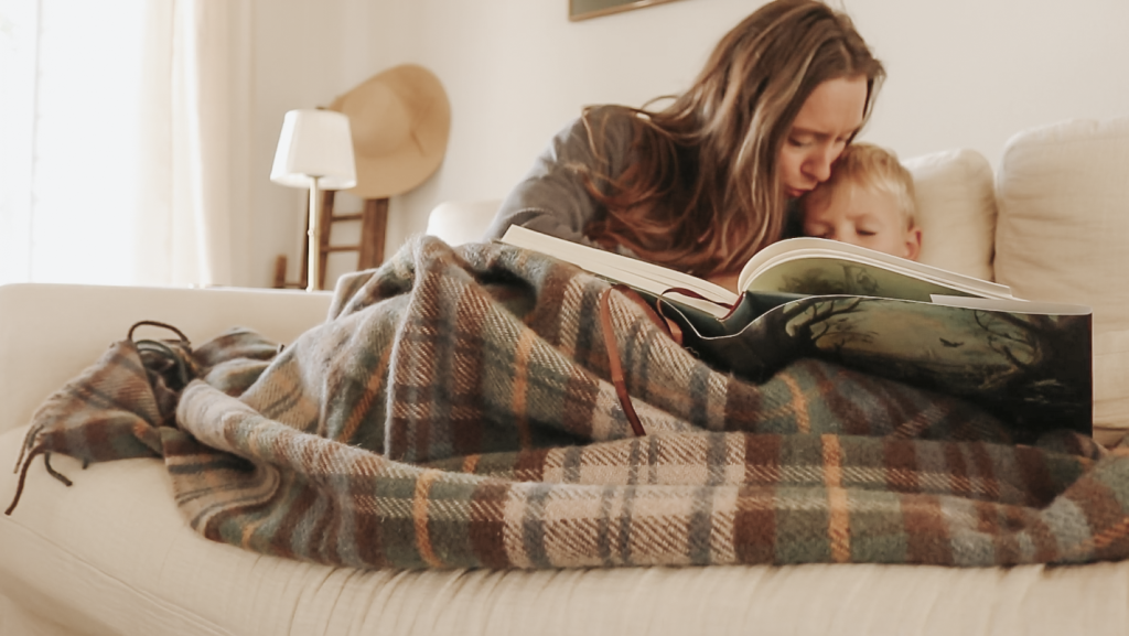 Christian homemaker sits on the couch reading a picture book to her child under a plaid blanket and giving him a kiss on the forehead.