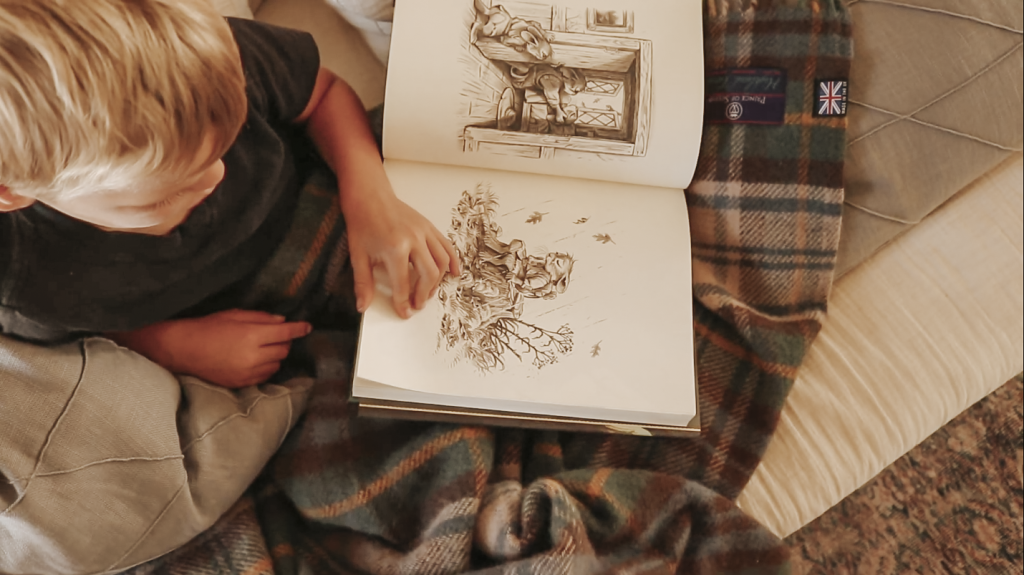 Young boy sitting on a couch under a plaid blanket, looking at and pointing to illustrations in an open book.