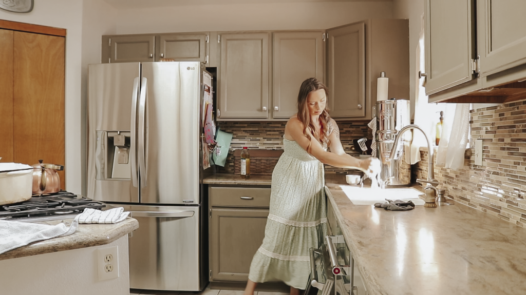 Christian homemaker wearing a light green dress stands at her kitchen sink wringing out a wet wash cloth.