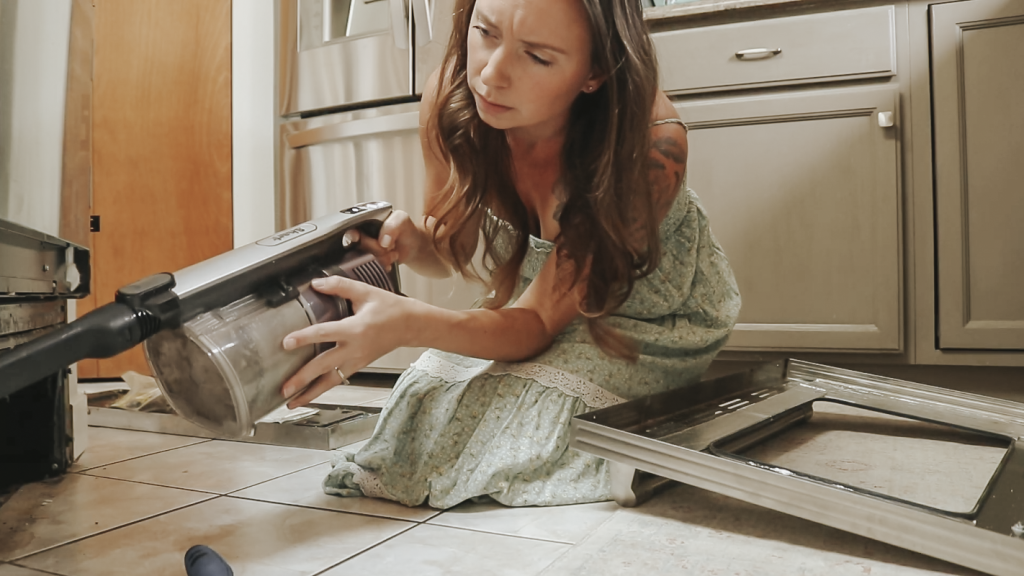 Christian homemaker wearing a light green dress is vacuuming under her cabinets in her kitchen.