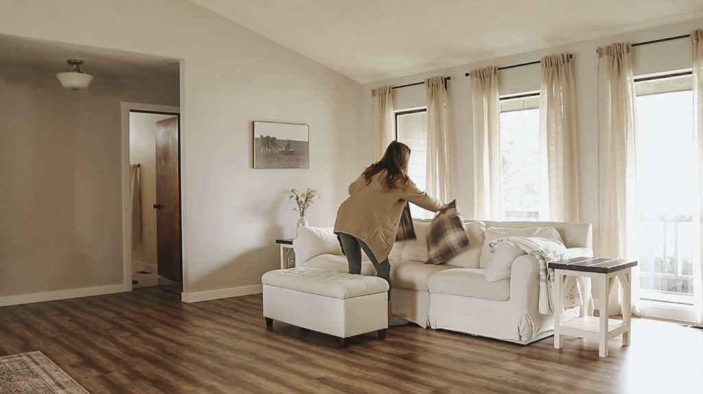Christian homemaker wearing jeans and a tan cardigan is arranging the pillows on a white couch in her living room.