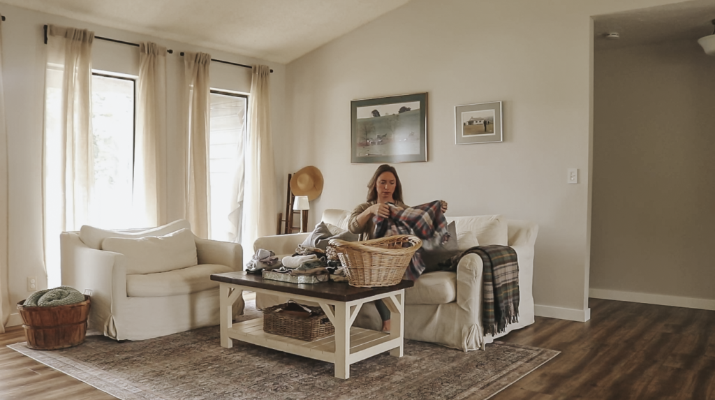 Christian homemaker wearing jeans and a tan cardigan is sitting on her couch in her living room folding a basket of laundry.