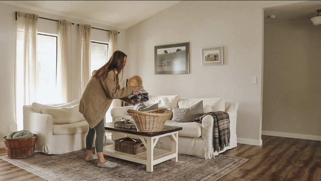 Christian homemaker wearing jeans and a tan cardigan places folded clothes into a laundry basket on the coffee table in her living room.