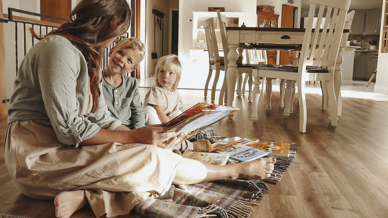Christian homemaker sits on the floor with her two children reading them a picture book. Her oldest child is looking at her and smiling.