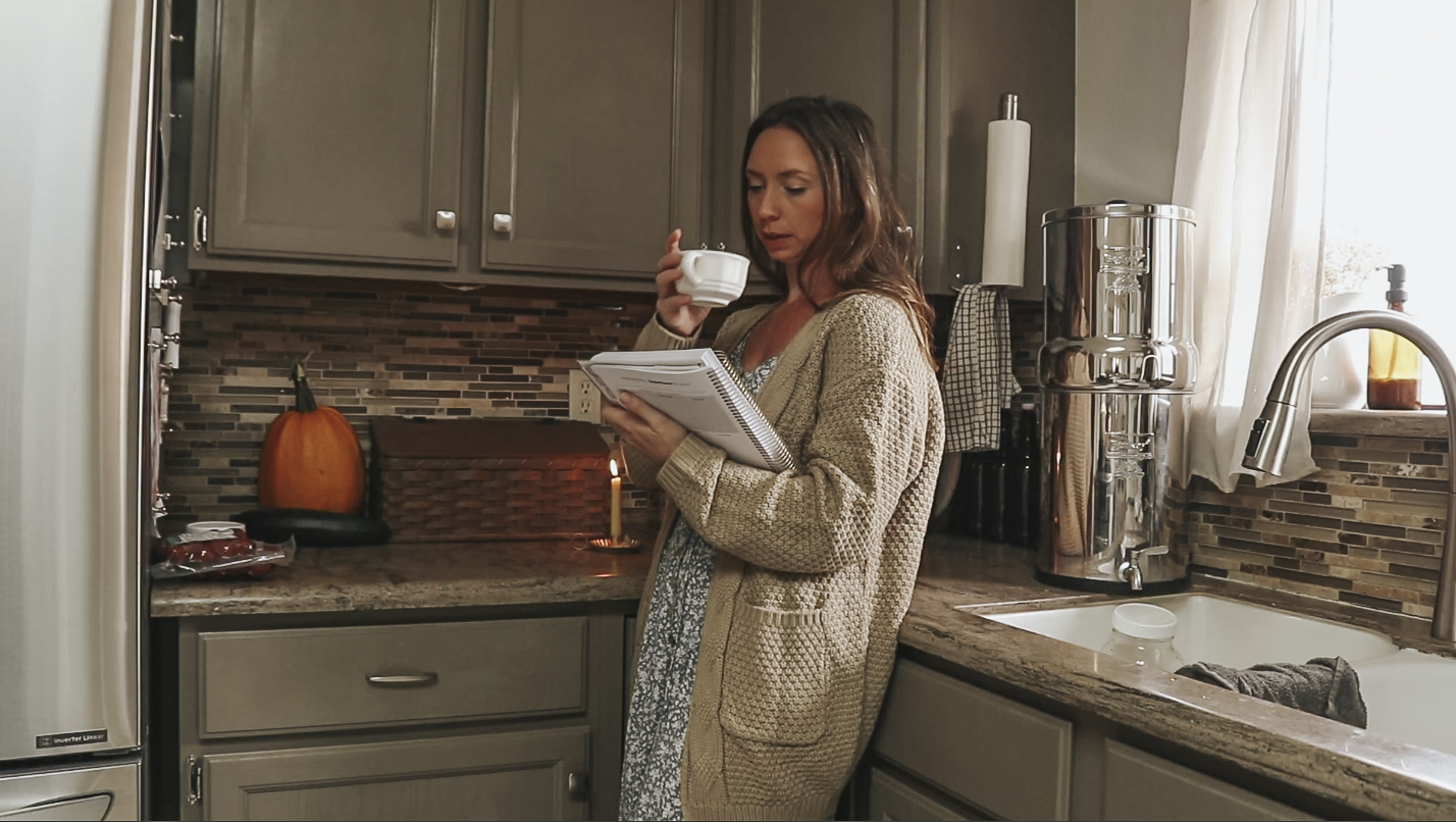 Christian homemaker wearing dress and cozy fall sweater leans up against her kitchen counter drinking a cup of coffee and reading a recipe book.