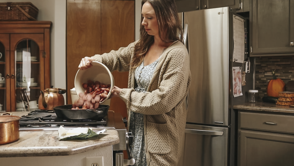 Christian homemaker wearing a blue floral dress and a tan cardigan pours cut up sausage from a bowl into a cast iron skillet.