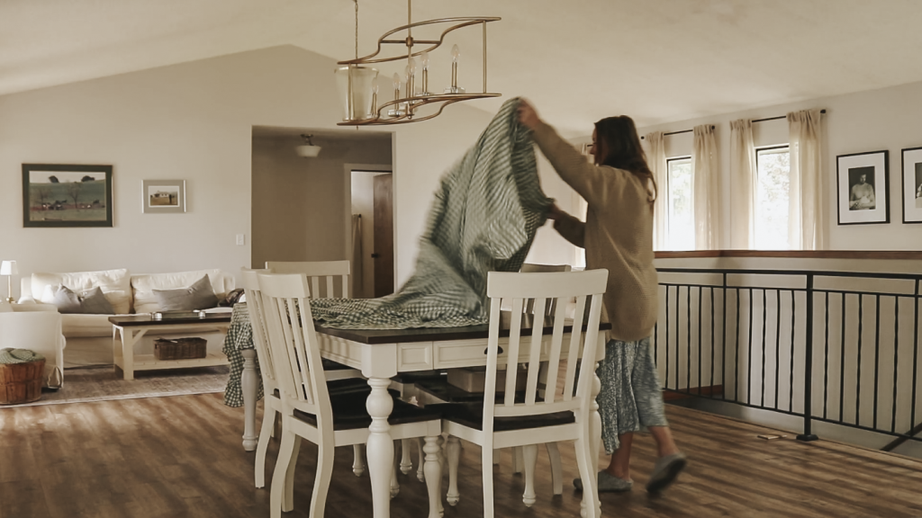 Christian homemaker wearing a blue floral dress and tan cardigan is spreading a table cloth over her kitchen table.