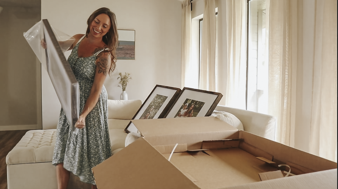 Christian homemaker wearing a green floral dress is unboxing a package with large printed photos in frames of her family.