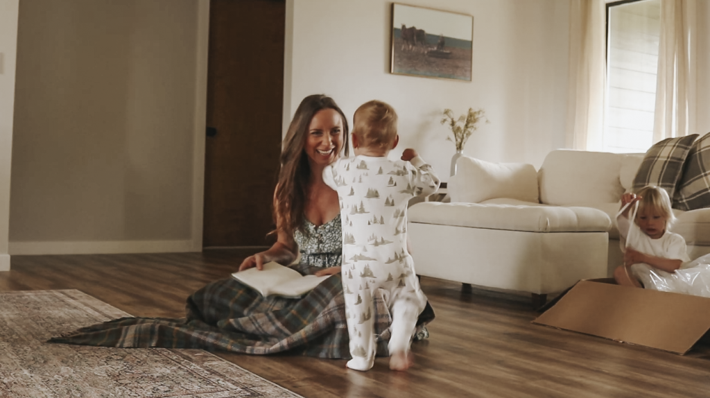 Christian homemaker wearing a floral dress and sitting on the floor with a blanket on her lap and book in her hand smiles at her toddler as he comes near to her.
