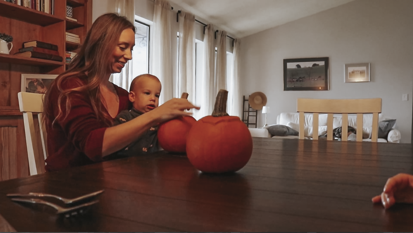 Christian homemaker sits at her kitchen table with her baby on her lap cutting into the top of a pumpkin.