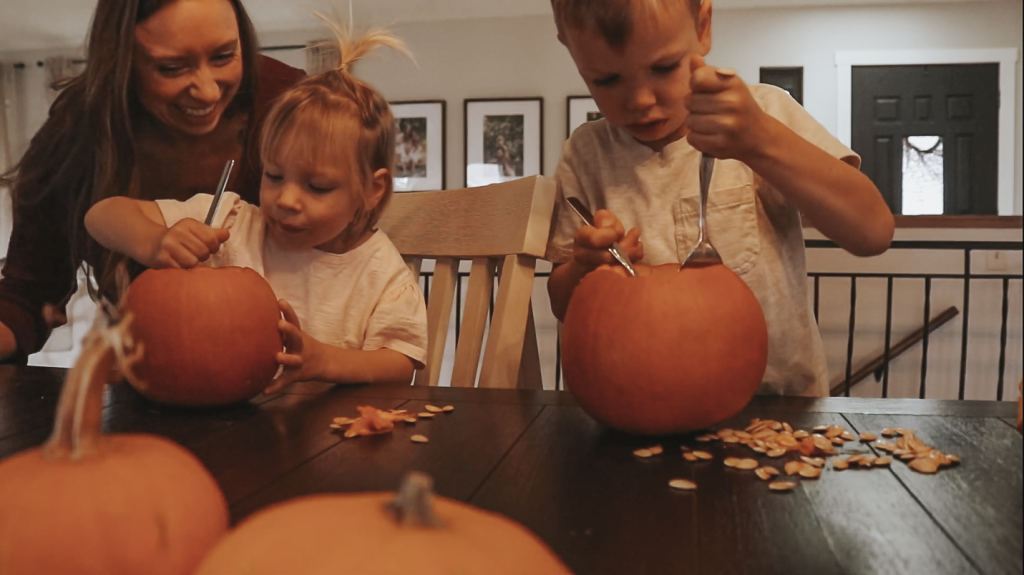 Christian homemaker sits at the kitchen table with her two young children as they work to scrape the seeds out of the inside of a pumpkin.