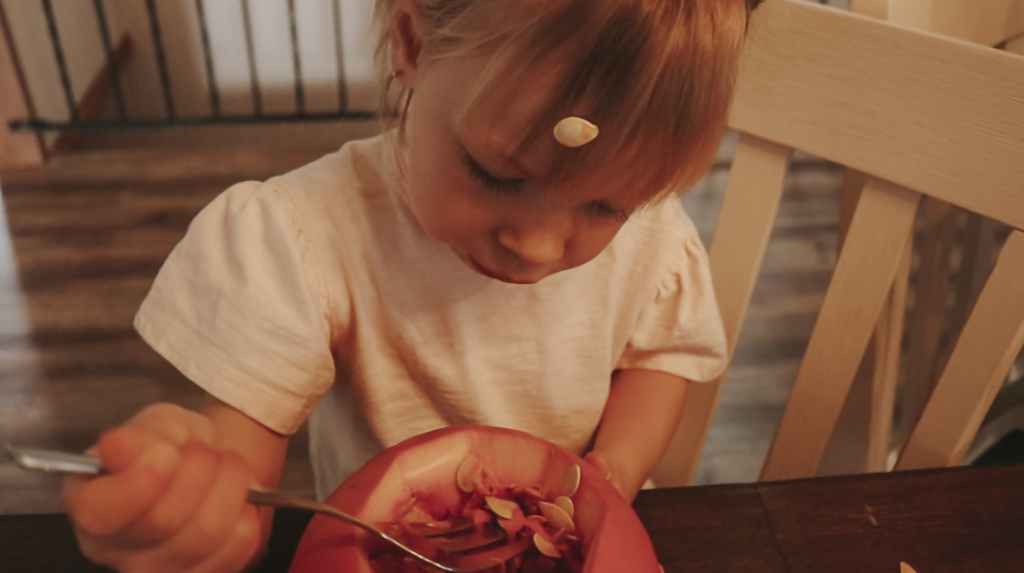 Christian homemaker's young child sitting at the kitchen with her pumpkin on the kitchen table and a pumpkin seed on her forehead.