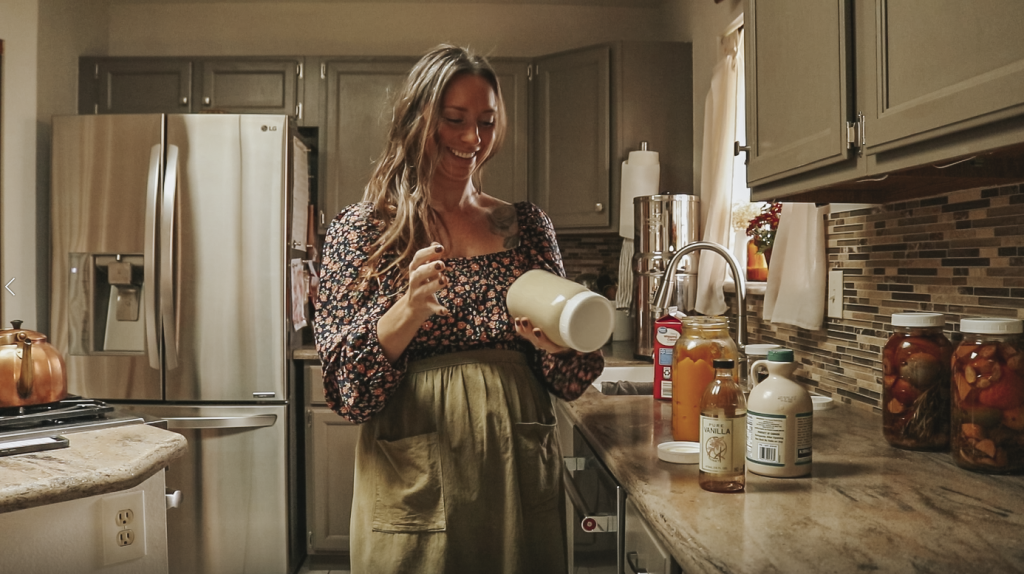 Christian homemaker wearing a black floral dress stands at her kitchen counter shaking her pumpkin spice coffee creamer in a glass mason jar with a lid.