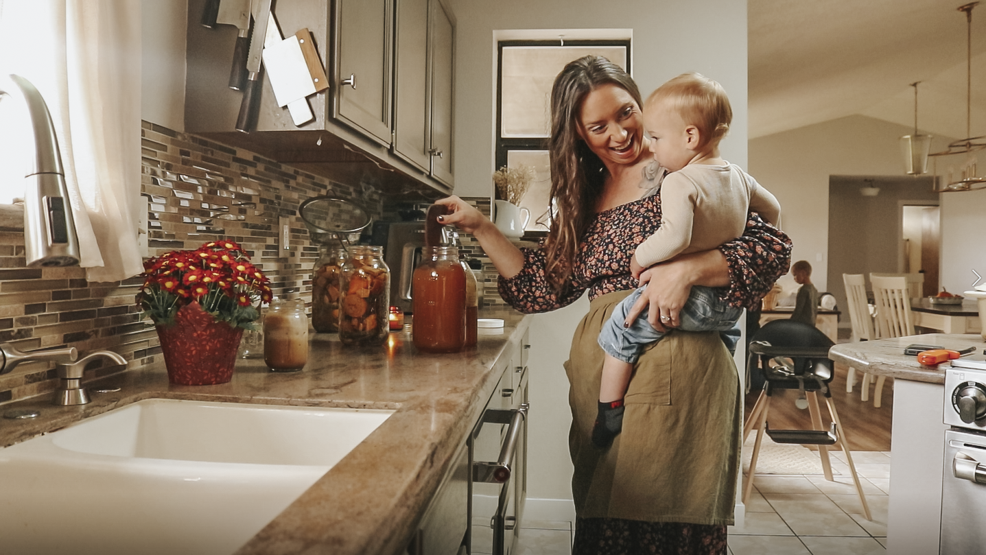 Christian homemaker wearing a brown floral dress stands at her counter holding her child and stirring her fire cider recipe.
