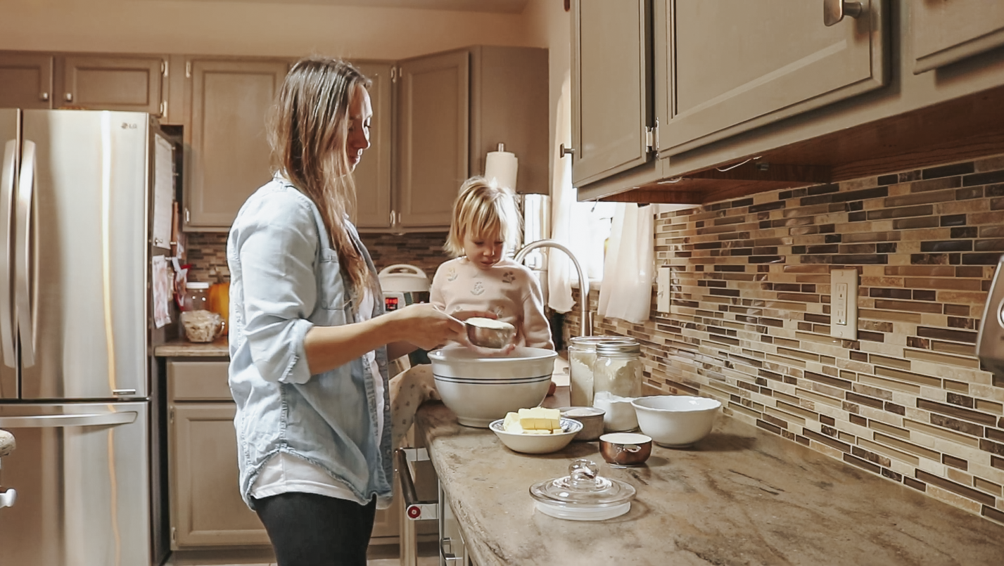 Christian mother and her young child standing together in a kitchen, mixing ingredients in a large bowl on the counter surrounded by other baking items.