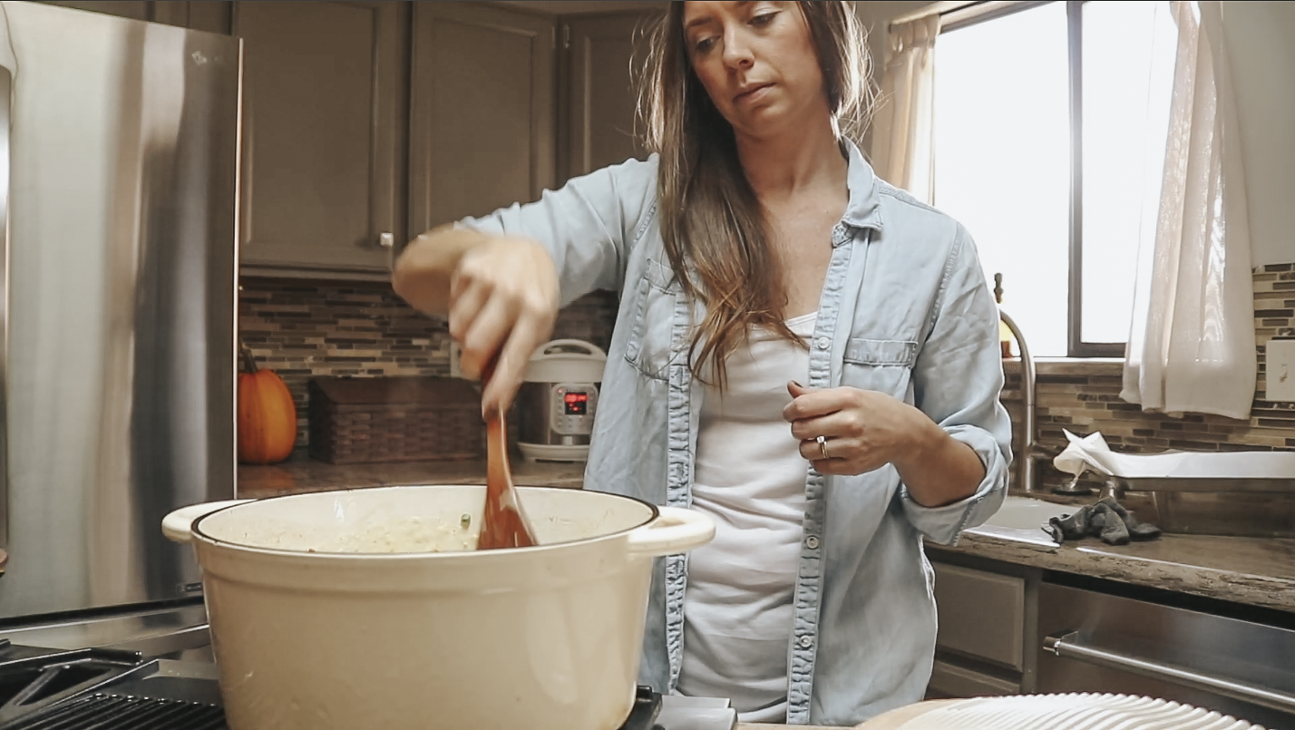 Christian homemaker wearing a white tank top, a denim button up top and leggings is standing at the stove stirring ingredients in a soup pot.
