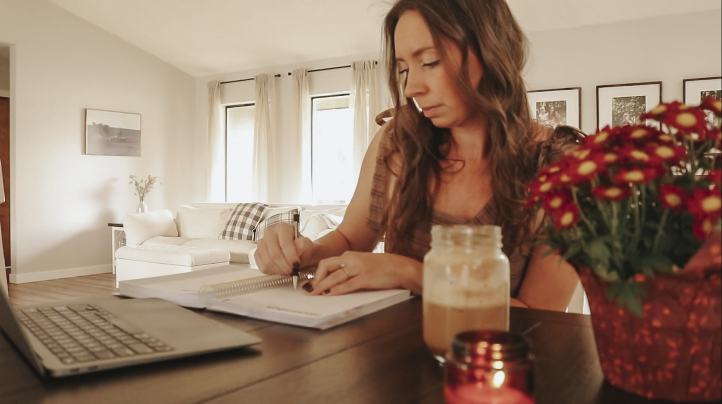 Christian homemaker is sitting at her kitchen table with her computer, planner and coffee in front of her and is writing things down in her planner.