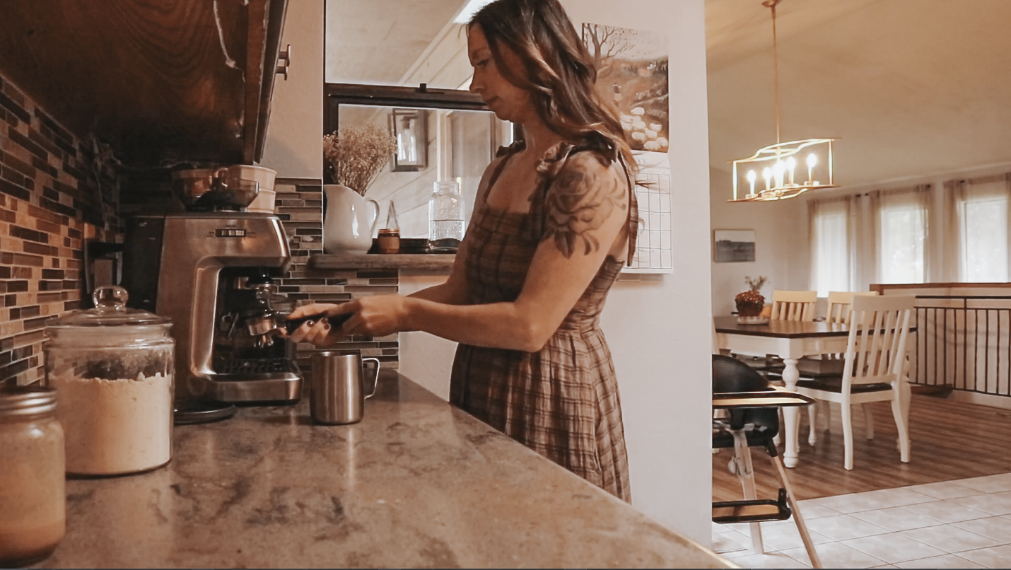 Christian homemaker wearing a brown plaid dress is standing at the counter in front of her coffee maker.