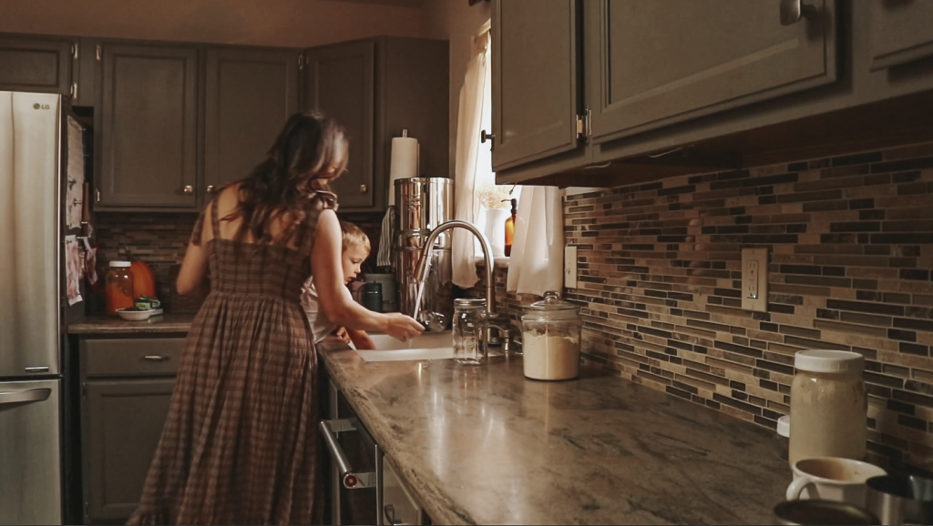Christian homemaker wearing a brown plaid dress is standing at the kitchen sink with her son and they are washing their hands.