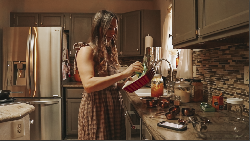 Christian homemaker wearing a brown plaid dress is greasing her glass bread pan with measuring cups and ingredients for making pumpkin bread on the kitchen counter in the background.
